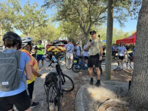 man speaking to a group of bikers in a park